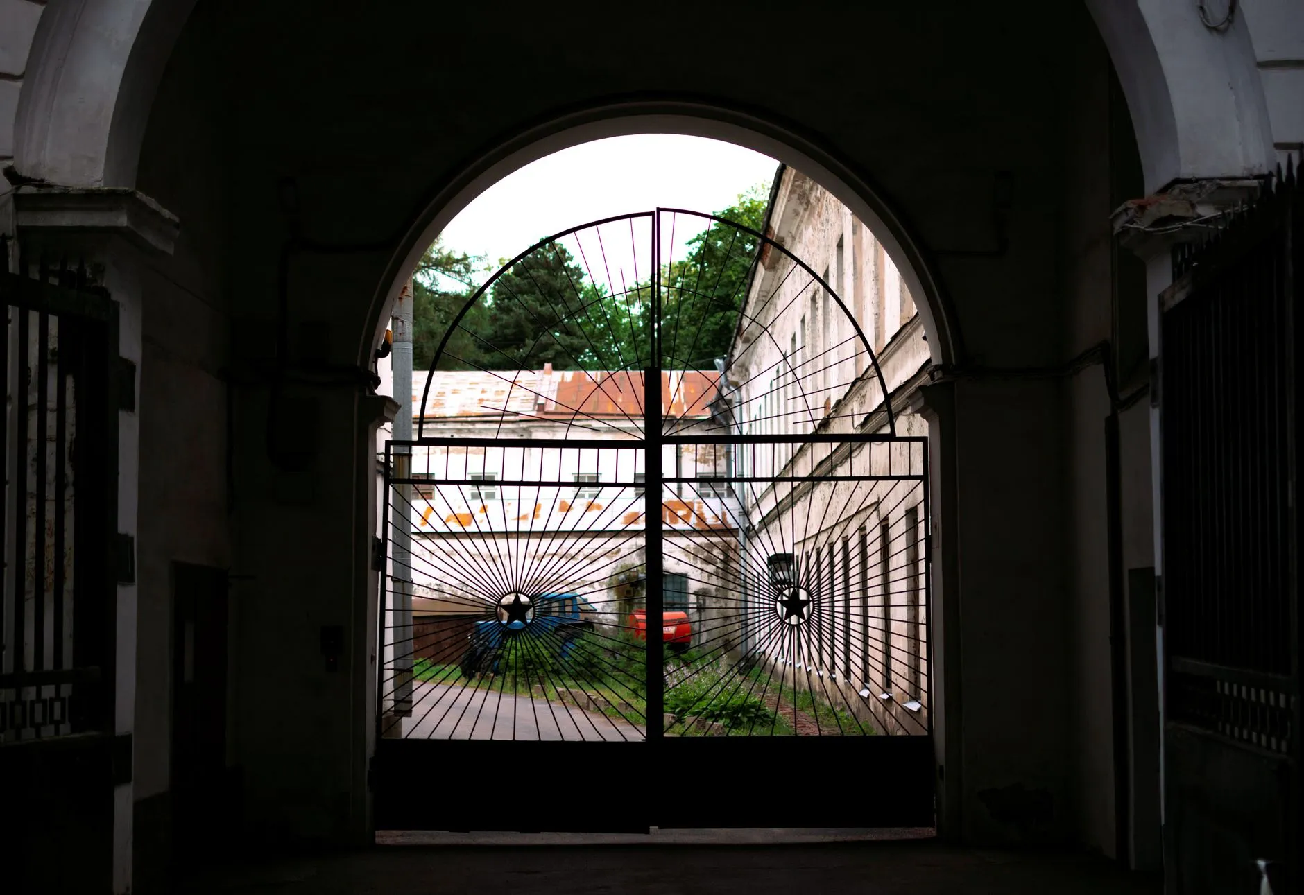 Portão de ferro em arco numa passagem escura, com um pátio iluminado ao fundo