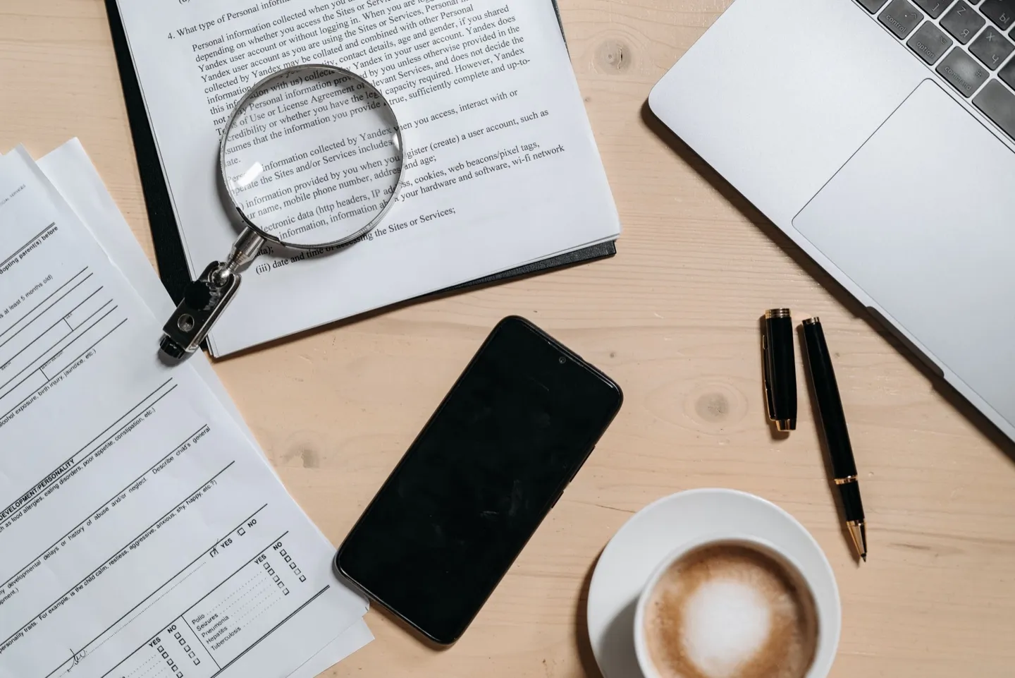 Documentos impressos sobre a mesa, com lupa, canetas e café ao lado de um notebook